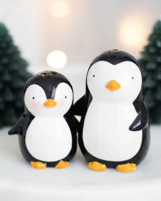 Front view of the hugging penguin salt and pepper shakers, with one adult and one baby penguin holding out their flippers for a hug, surrounded by festive fairy lights and miniature Christmas trees.