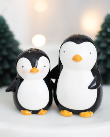 Front view of the hugging penguin salt and pepper shakers, with one adult and one baby penguin holding out their flippers for a hug, surrounded by festive fairy lights and miniature Christmas trees.