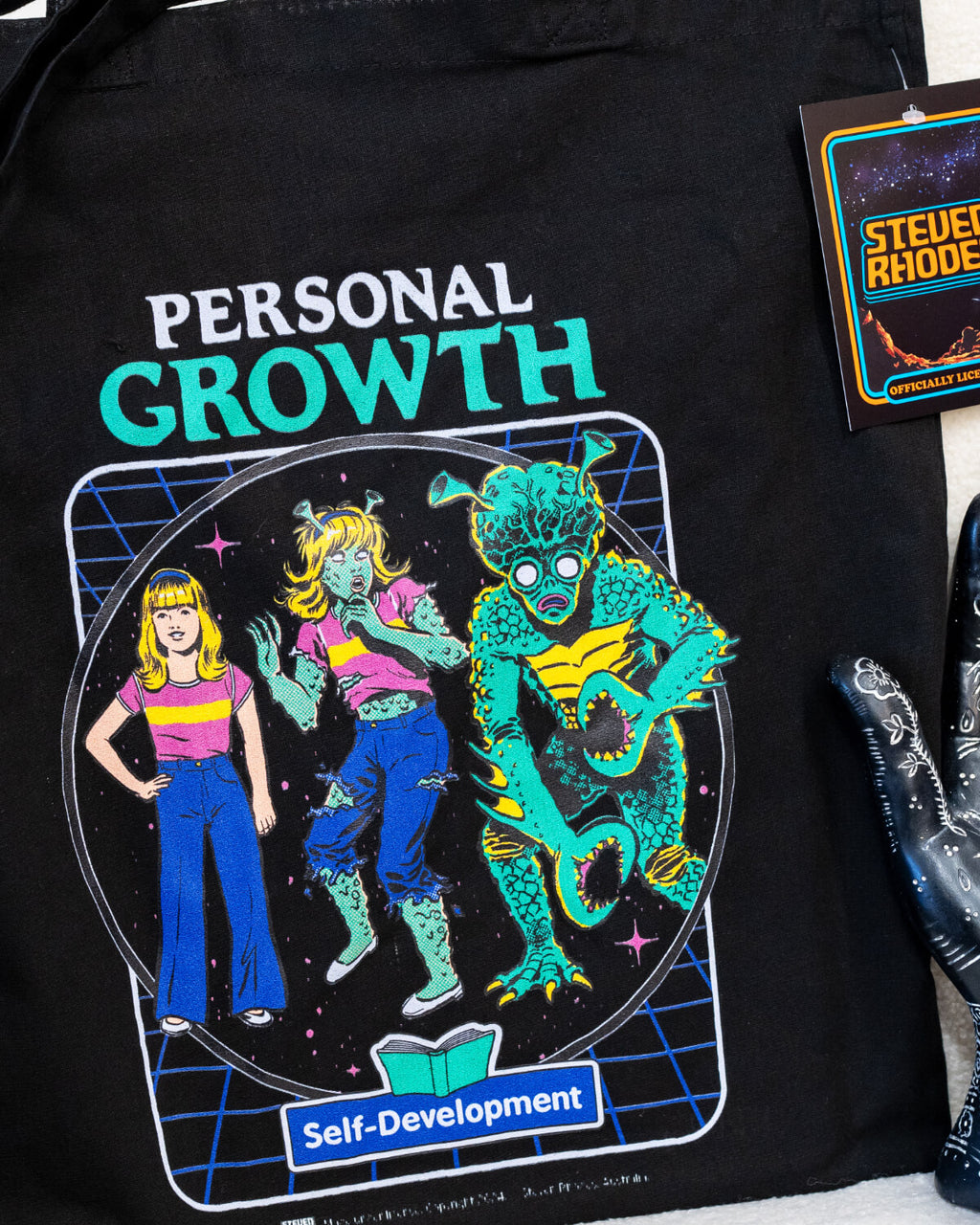 A black tote bag featuring Steven Rhodes’ “Personal Growth” design, showcasing a retro-inspired self-development illustration with vibrant neon colours and a humorous theme. The tote includes long handles for easy carrying and is labelled as officially licensed merchandise. Perfect for shopping, work, or travel.