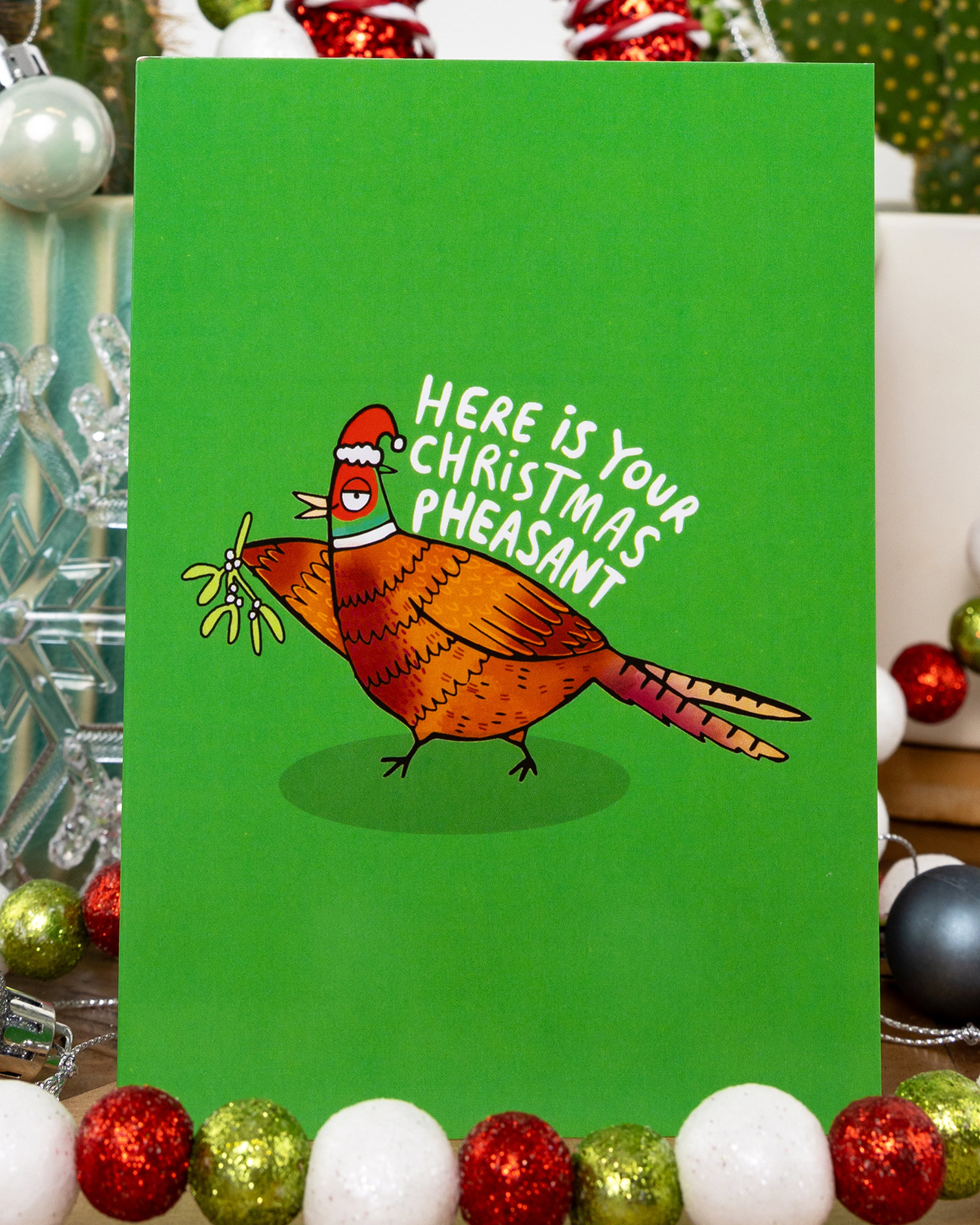 A bright green Christmas card by Rebel Romance featuring a whimsical illustration by Katie Abey of a colourful pheasant wearing a Santa hat, holding mistletoe. The card reads “Here is your Christmas Pheasant” in white lettering above the bird. Surrounded by festive baubles and decorations, the design adds fun and charm to holiday greetings. A quirky card perfect for animal lovers and fans of alternative style.