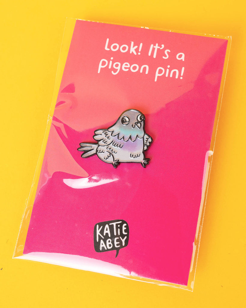 The pigeon enamel pin displayed on a bright pink Katie Abey backing card that reads “Look! It’s a pigeon pin!” against a yellow background.