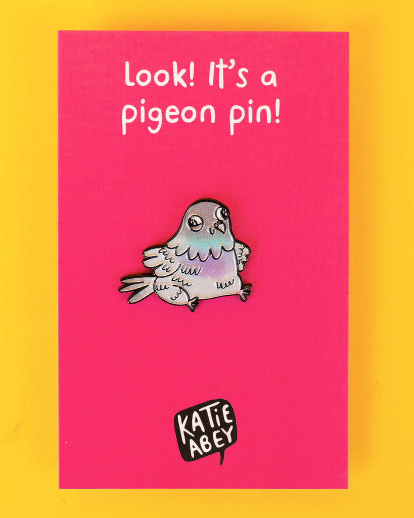 The pigeon pin on a bright pink backing card with Katie Abey branding, photographed flat against a yellow background.