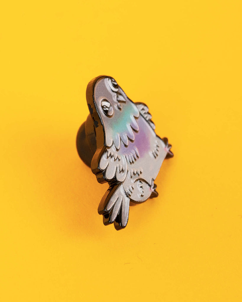 Close-up of the Katie Abey pigeon enamel pin showing a chonky pastel pigeon with cute cartoon eyes, photographed on a vibrant yellow background.