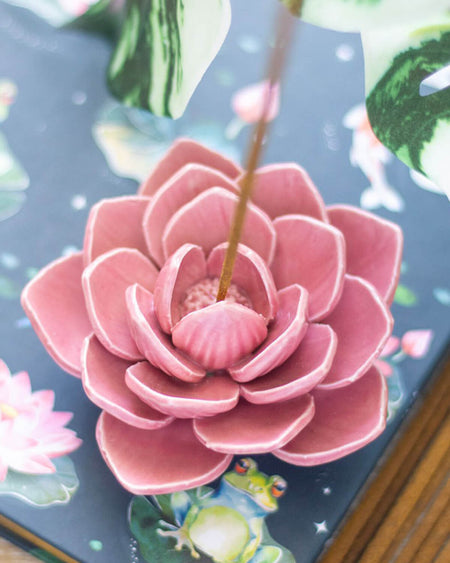 A pink water lily incense holder with a delicately sculpted resin lotus flower, securely holding an incense stick on a dreamy, floral-patterned surface