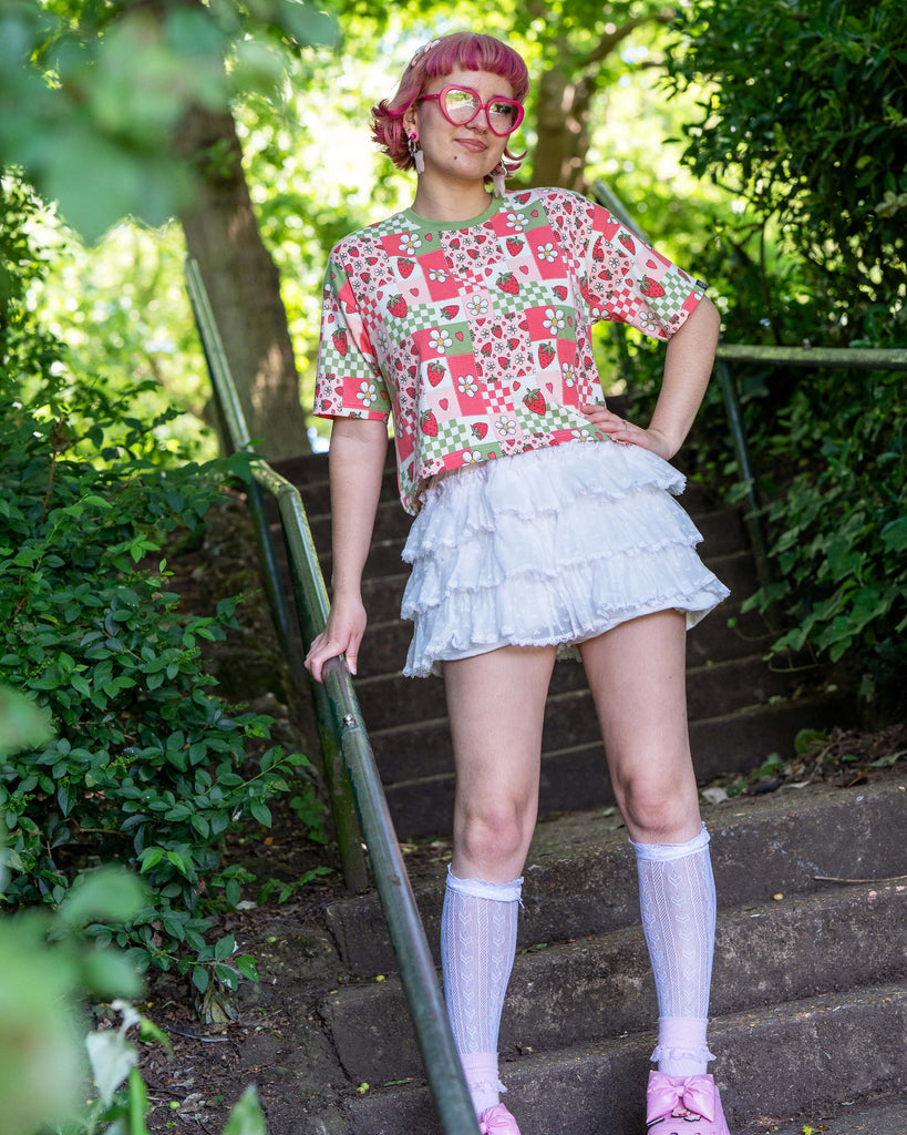 Jess, a pink haired model with heart shaped glasses wearing the strawberry picnic crop top with white ruffle shorts, white knee high socks and pink platform crocs. She is stood on a park staircase with green foliage all around her. The strawberry picnic print is a sage green, pastel red, baby pink and white checkerboard with strawberries and flower blossoms all over.