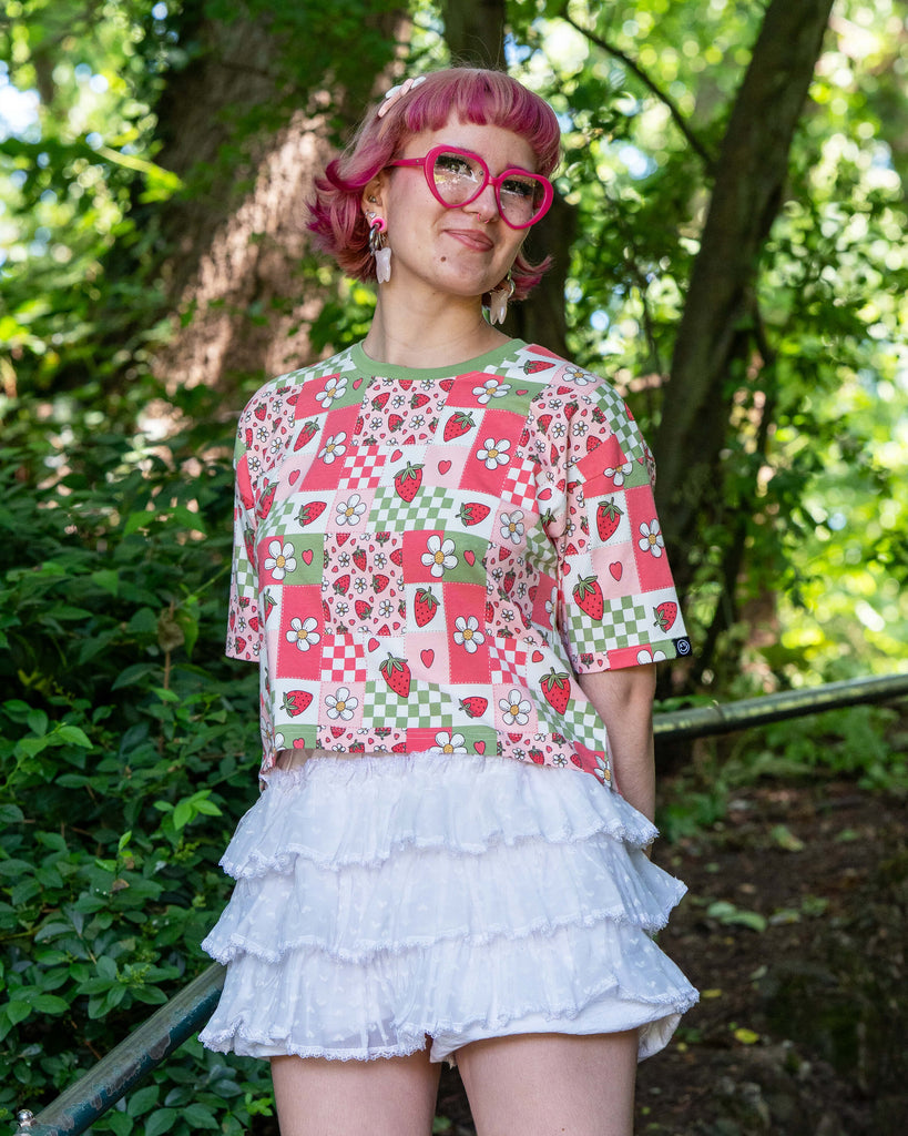 Jess, a pink haired model with heart shaped glasses wearing the strawberry picnic crop top with white ruffle shorts,. She is stood on a park staircase smiling with green foliage all around her. The strawberry picnic print is a sage green, pastel red, baby pink and white checkerboard with strawberries and flower blossoms all over.