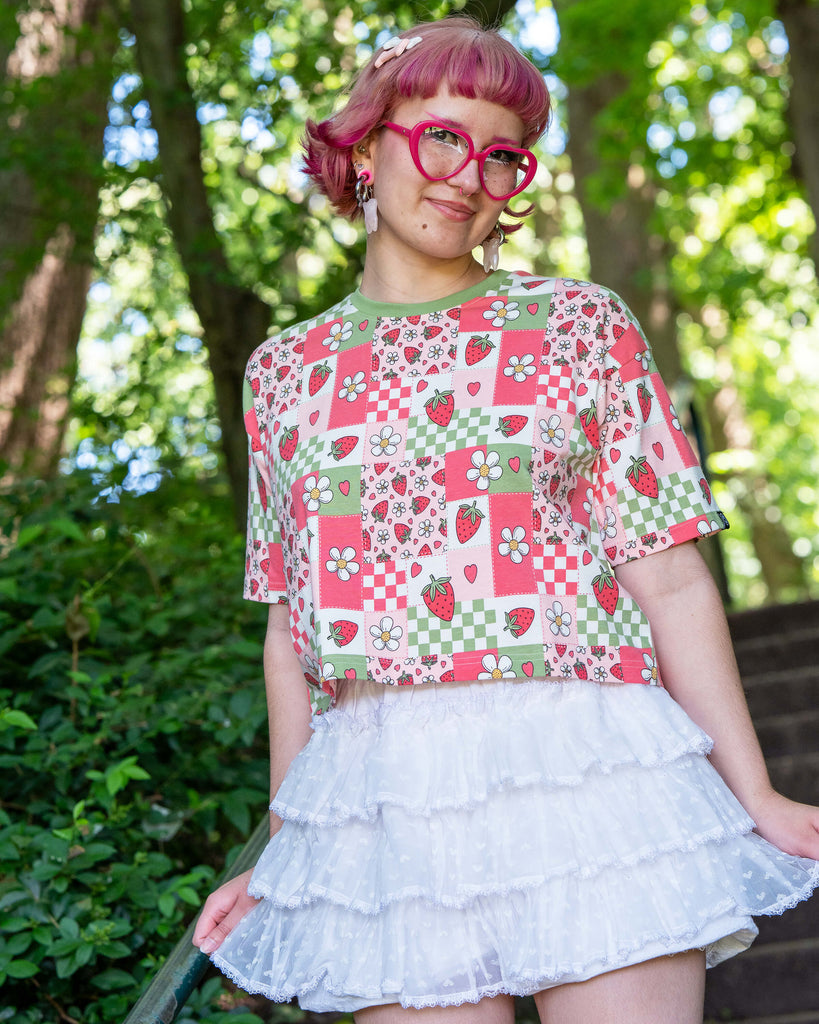 Jess, a pink haired model with heart shaped glasses wearing the strawberry picnic crop top with white ruffle shorts. She is stood on a park staircase with green foliage all around her. The strawberry picnic print is a sage green, pastel red, baby pink and white checkerboard with strawberries and flower blossoms all over.