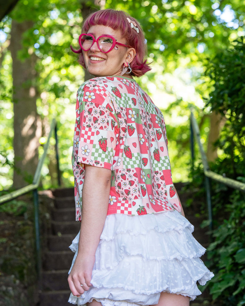 Jess, a pink haired model with heart shaped glasses wearing the strawberry picnic crop top with white ruffle shorts. She is stood on a park staircase with green foliage all around her facing away and smiling over her shoulder to camera. The strawberry picnic print is a sage green, pastel red, baby pink and white checkerboard with strawberries and flower blossoms all over.