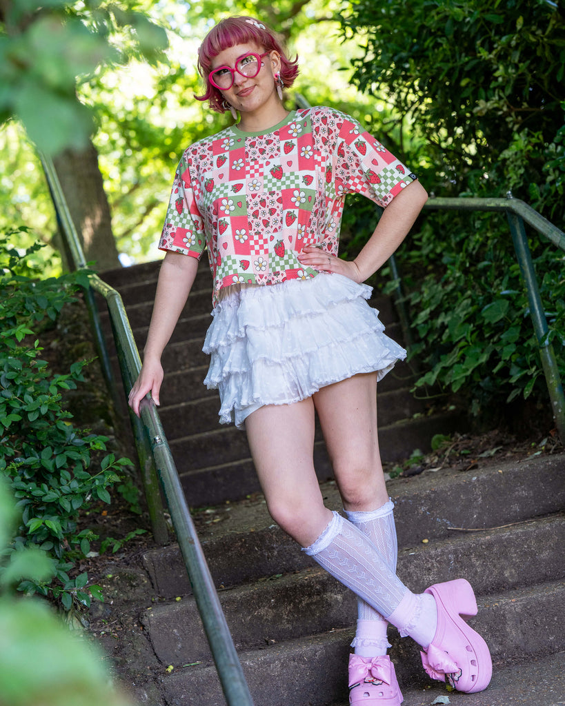 Jess, a pink haired model with heart shaped glasses wearing the strawberry picnic crop top with white ruffle shorts, white knee high socks and pink platform crocs. She is stood on a park staircase with green foliage all around her. The strawberry picnic print is a sage green, pastel red, baby pink and white checkerboard with strawberries and flower blossoms all over.