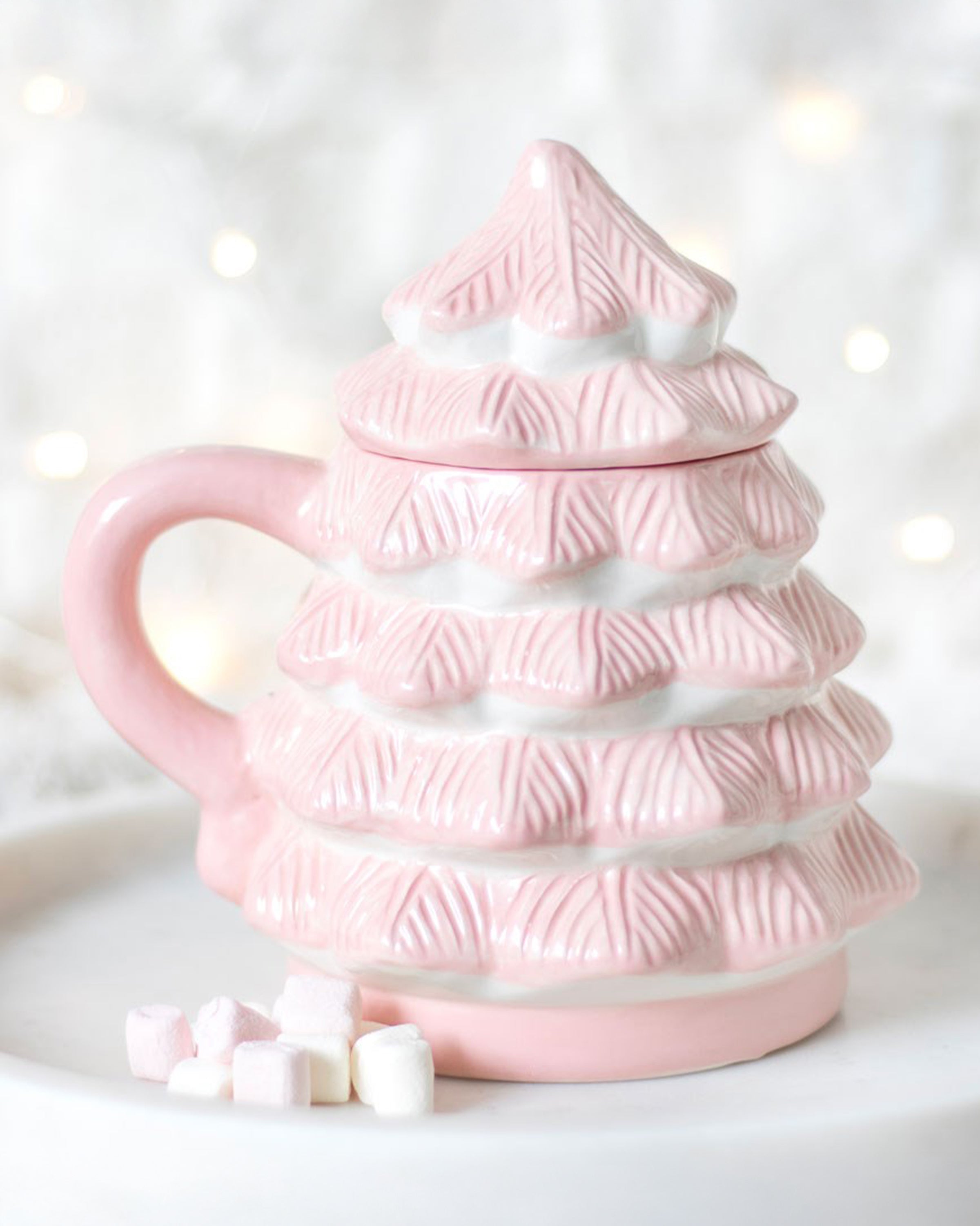 Aesthetic photo of the Rebel Romance Pink Christmas Tree Mug styled on a white tray with mini marshmallows. The pastel pink ceramic mug is shaped like a Christmas tree with a ridged, snowy design and a removable lid. A cosy winter backdrop with fairy lights completes the festive vibe.