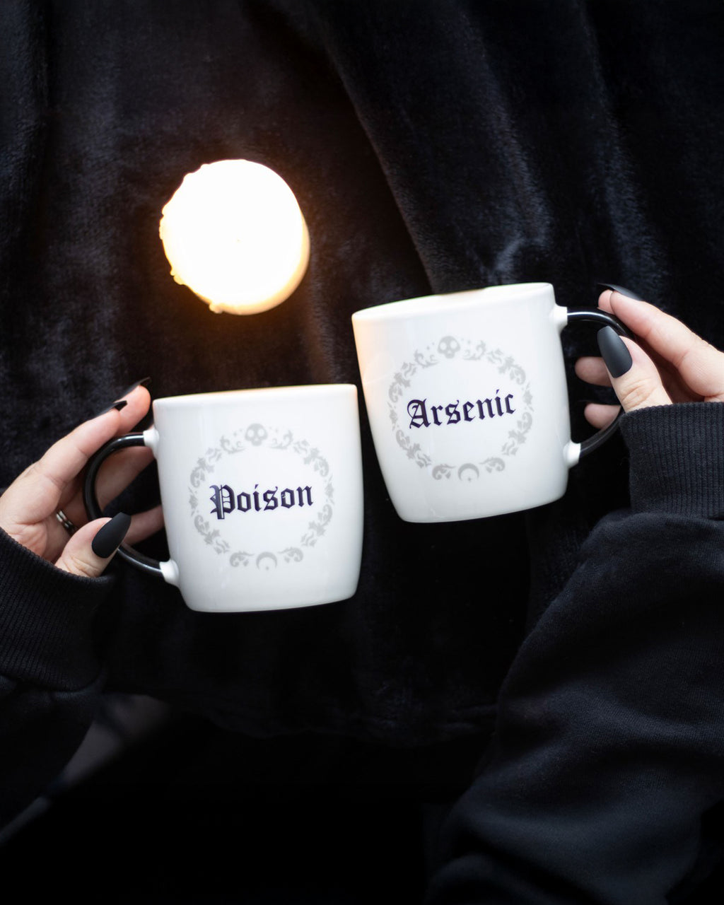 A moody lifestyle shot of the Poison and Arsenic Mugs being held over a black velvet background, with gothic black nail polish and candlelight adding to the eerie, witchy aesthetic.