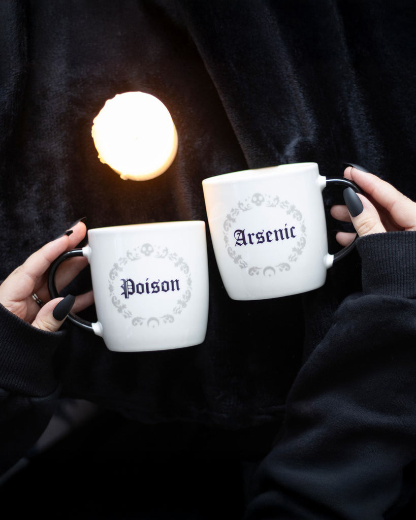 A moody lifestyle shot of the Poison and Arsenic Mugs being held over a black velvet background, with gothic black nail polish and candlelight adding to the eerie, witchy aesthetic.