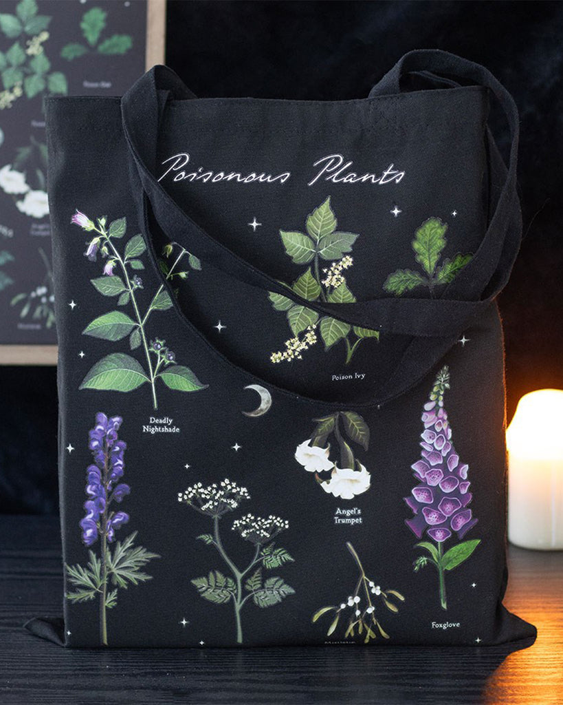 Poisonous Plants Polycotton Tote Bag by Rebel Romance styled with candlelight, showcasing detailed illustrations of toxic plants on a black background. A gothic-inspired eco tote for witchy vibes and dark botanical fashion lovers.