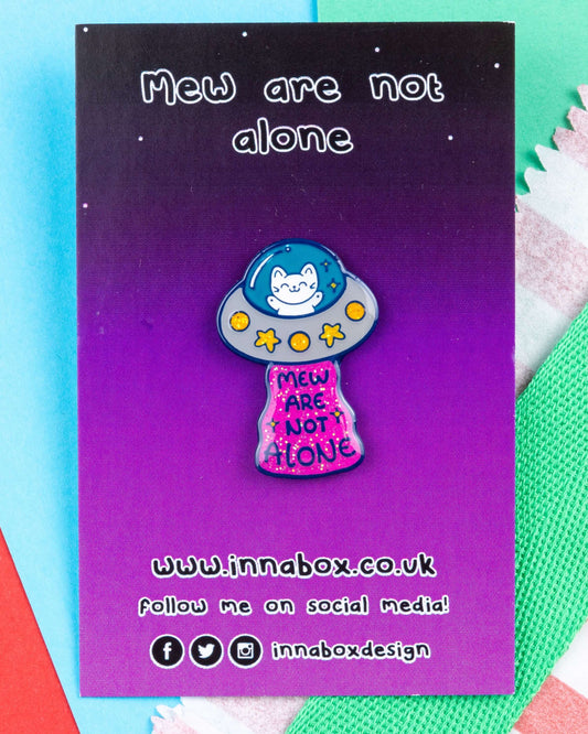 Glittery enamel pin of a cat in a UFO reading "Mew Are Not Alone", placed on a bright blue background with colourful paper and fabric details. Created by Innabox and sold by Rebel Romance, a small UK brand celebrating mental health awareness through quirky, alternative accessories.