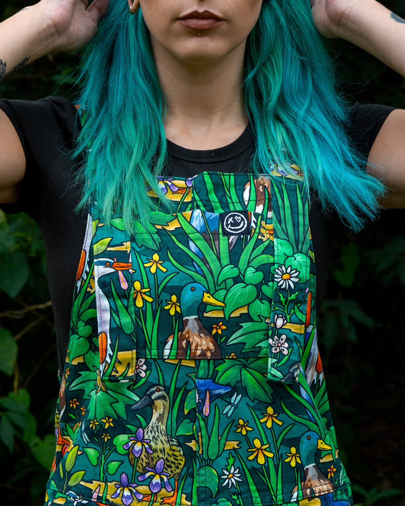 Detail shot of the Rebel Romance Pond Birds Dungarees bib pocket, showing a colourful nature-inspired print with mallard ducks, herons, daisies, and water plants. Made from eco-friendly stretch twill with adjustable straps, designed for comfort and playful everyday wear.