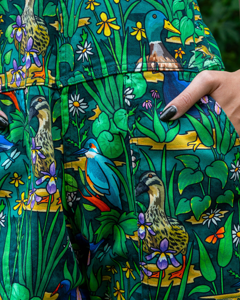Close-up of the Pond Birds dungarees pocket, decorated with kingfishers, ducks, and wildflowers in a detailed green wetland print. Ethically crafted stretch twill dungarees with roomy pockets and inclusive fit, from the UK indie brand Rebel Romance.