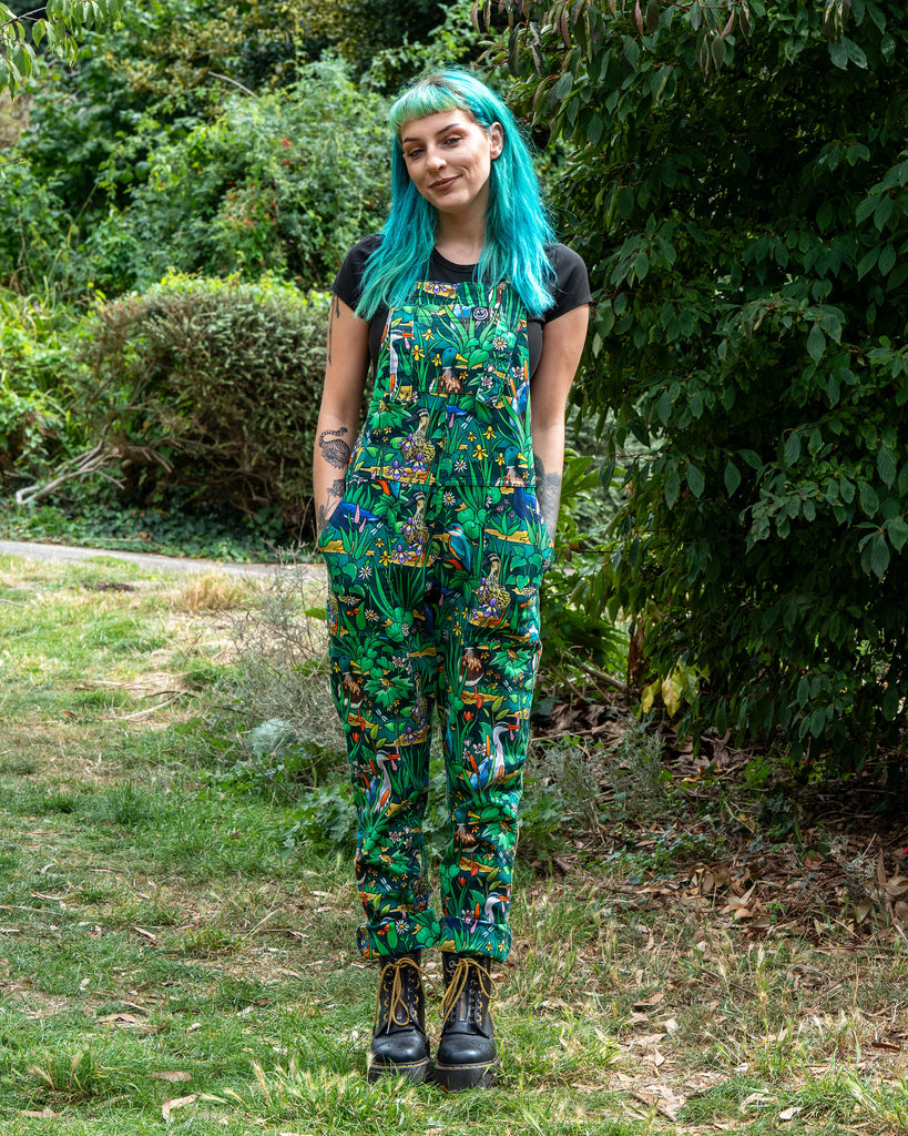 Claudia wears the Rebel Romance Pond Birds Stretch Dungarees in a lush outdoor setting. The bold nature print features ducks, herons, and flowers on green twill fabric. Ethically made dungarees with adjustable straps, roll-up hems, and pockets.