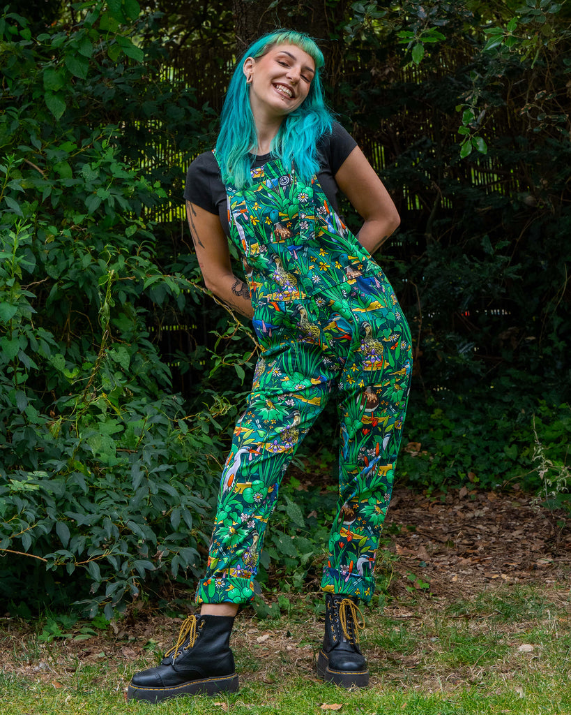 Claudia smiles while modelling the Rebel Romance Pond Birds Dungarees with a lively green print of ducks, herons, and pond plants. Ethically made dungarees with side and bib pockets, roll-up hems, and adjustable straps.