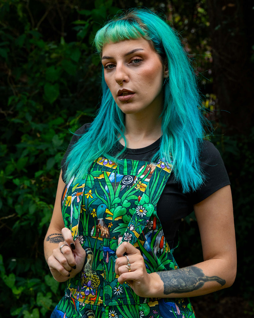 Close-up of Claudia adjusting the straps of the Rebel Romance Pond Birds Dungarees, highlighting the colourful duck and flower print on green twill fabric. A bold, eco-friendly slow fashion piece by Rebel Romance.