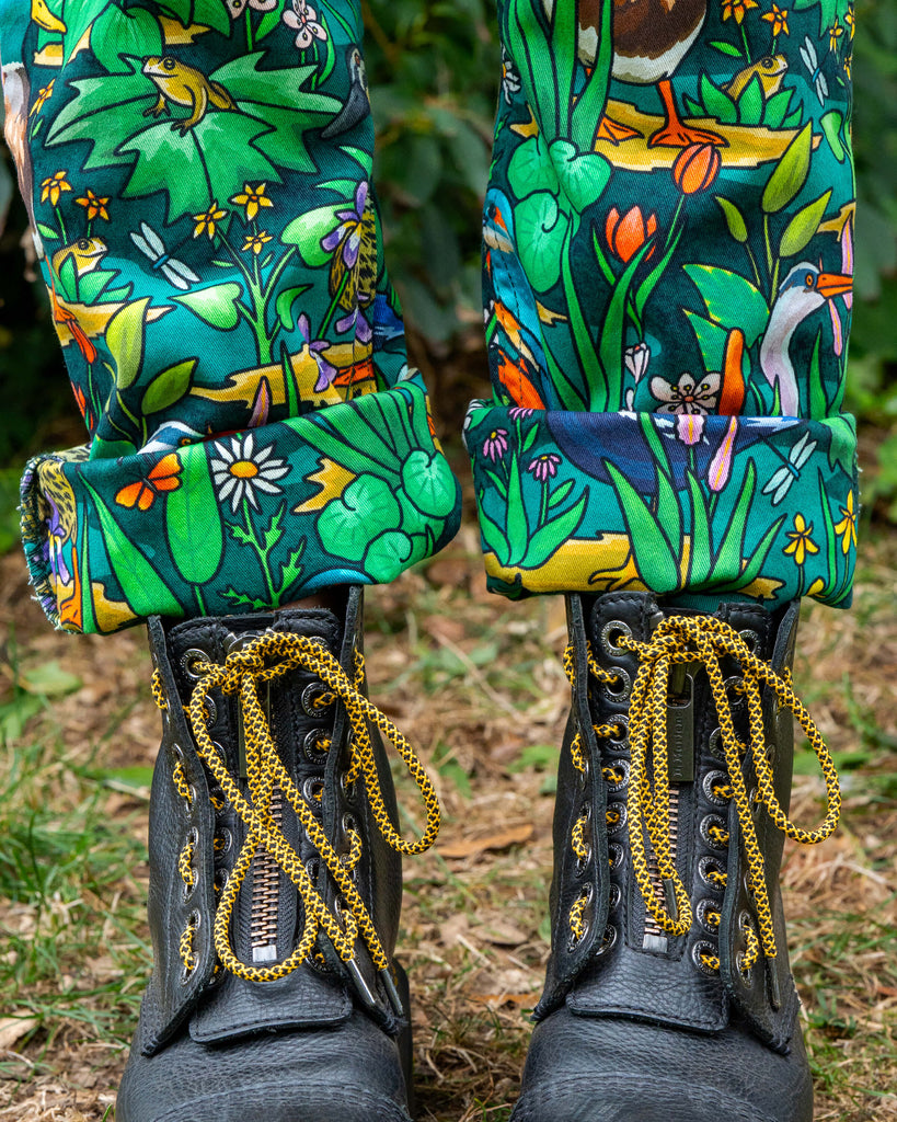 Close-up of the Rebel Romance Pond Birds Dungarees rolled up legs, showing intricate pond life illustrations including frogs, ducks, and wildflowers. Worn with chunky black boots for an alternative fashion look.