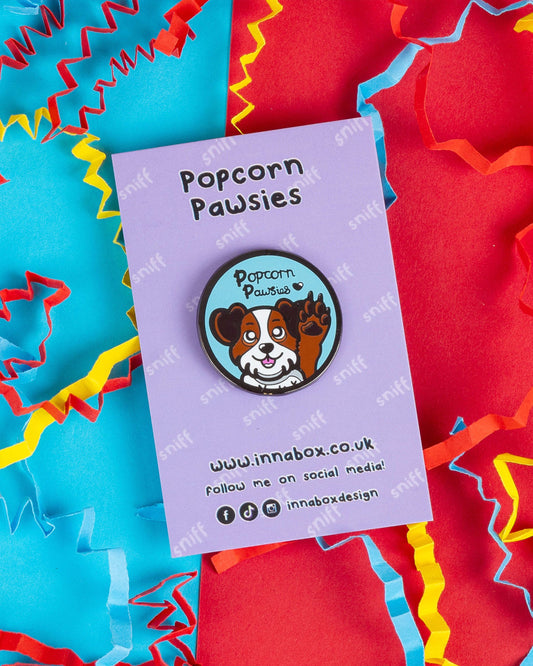 Popcorn Pawsies enamel pin on a pastel purple backing card with “Popcorn Pawsies” written above. The pin shows a smiling cartoon dog with a raised paw, resembling popcorn toe beans. Surrounded by colourful red, yellow and blue confetti. Designed by Innabox and sold by Rebel Romance, a UK small biz focused on alternative fashion and gifts.