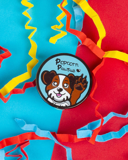 Popcorn Pawsies round enamel pin on a bright red and turquoise background with colourful paper confetti. The pin features a cute brown and white cartoon dog holding up a paw with the words “Popcorn Pawsies” above. A playful design by Innabox, sold by Rebel Romance – a UK-based small business that celebrates alternative style and quirky gifts.