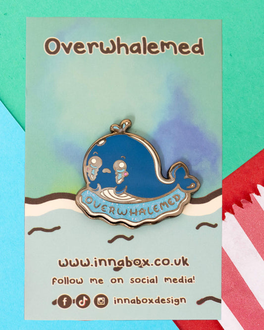 Enamel pin on a backing card that reads "Overwhalemed" featuring a blue glittery whale with teary eyes and the word “OVERWHALEMED” beneath. Surrounded by colourful paper decorations. Product designed by Innabox and stocked by Rebel Romance, a small UK business known for alternative fashion and quirky gifts