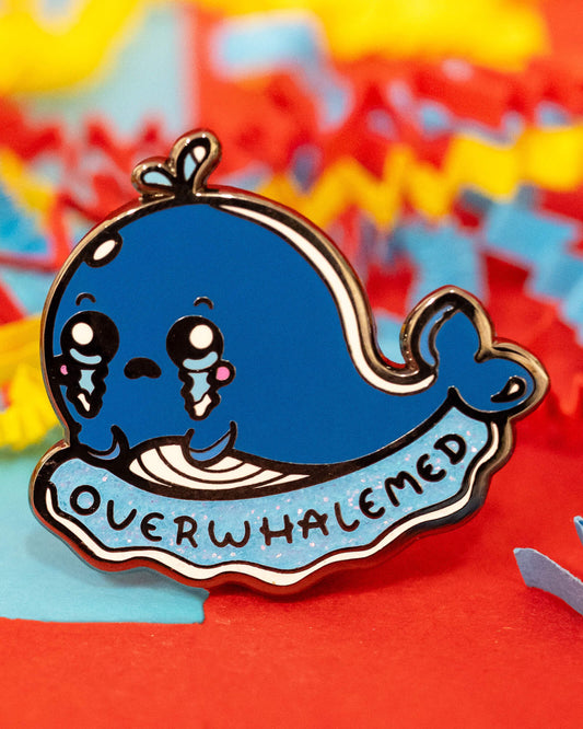 Close-up of the Overwhalemed enamel pin by Rebel Romance, showing a glittery blue whale with a sad expression and text reading “OVERWHALEMED.” Set against a colourful background with red, blue, and yellow zig-zag paper strips. A playful and comforting pin designed by Innabox.