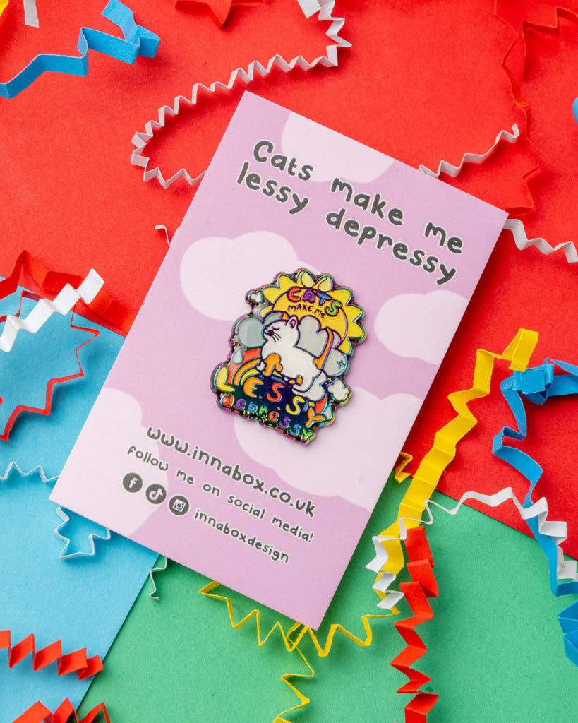A colourful enamel pin on a backing card that reads "Cats make me lessy depressy." The pin features a white cartoon cat with a rainbow and cloud background, surrounded by cheerful shapes and colours. The pin is displayed on a pink and purple polka dot card, surrounded by multicoloured paper confetti. From Rebel Romance, a small UK business selling alternative gifts and fashion for millennials.
