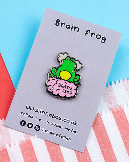 Brain Frog enamel pin displayed on a lilac backing card with the product name above it. The design features a green cartoon frog sitting on a pink brain with the words “Brain Frog”, representing brain fog. Set against a colourful red and blue background with tissue paper. Designed by Innabox and sold by Rebel Romance, a UK indie brand focused on unique gifts and alternative style.