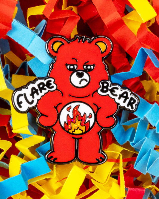 Close-up of the Flare Bear enamel pin showing a red bear with a tired expression and a glittery fire belly symbol. The bear’s paws say "Flare" and "Bear" in bold text. Laid on a colourful background of shredded red, yellow, and blue paper. Designed by Innabox and available at Rebel Romance, a UK alternative fashion and gift brand.