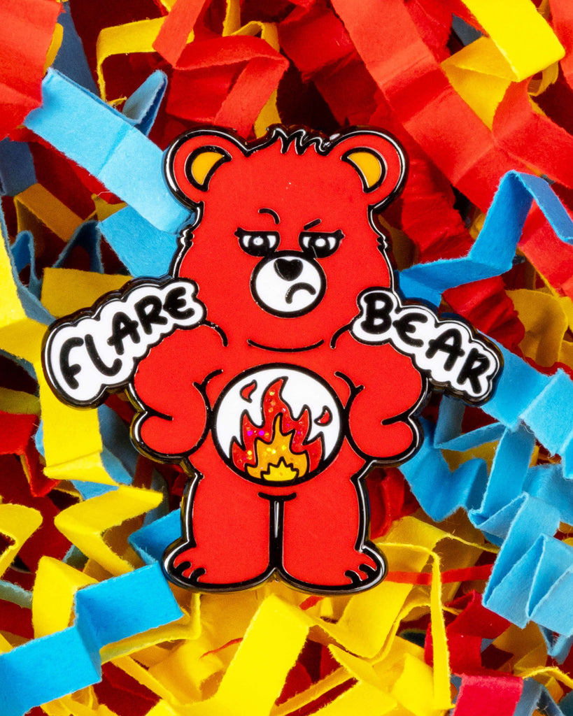 Close-up of the Flare Bear enamel pin showing a red bear with a tired expression and a glittery fire belly symbol. The bear’s paws say "Flare" and "Bear" in bold text. Laid on a colourful background of shredded red, yellow, and blue paper. Designed by Innabox and available at Rebel Romance, a UK alternative fashion and gift brand.