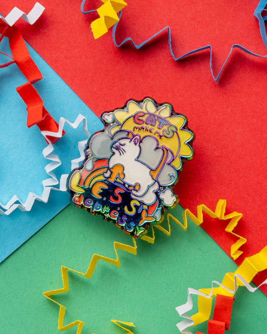 Close-up of the “Cats Make Me Lessy Depressy” enamel pin, showing a white cartoon cat, bold rainbow lettering, and fun background shapes. The pin has rainbow metal plating and pink rubber backs. Photographed on a colourful flat lay with red, blue, and green sections and paper confetti. Sold by Rebel Romance, an alternative UK gift and fashion brand.