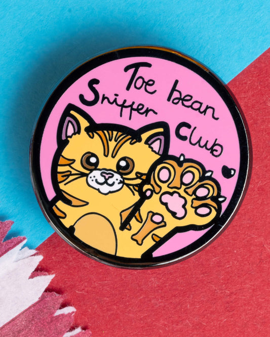 Close-up of the Toe Bean Sniffer Club enamel pin showing a smiling orange cat and raised paw with pink toe beans. Text reads "Toe Bean Sniffer Club" on a pastel pink background. Ideal for cat lovers and collectors. Sold by Rebel Romance, a UK alternative fashion and gift shop.