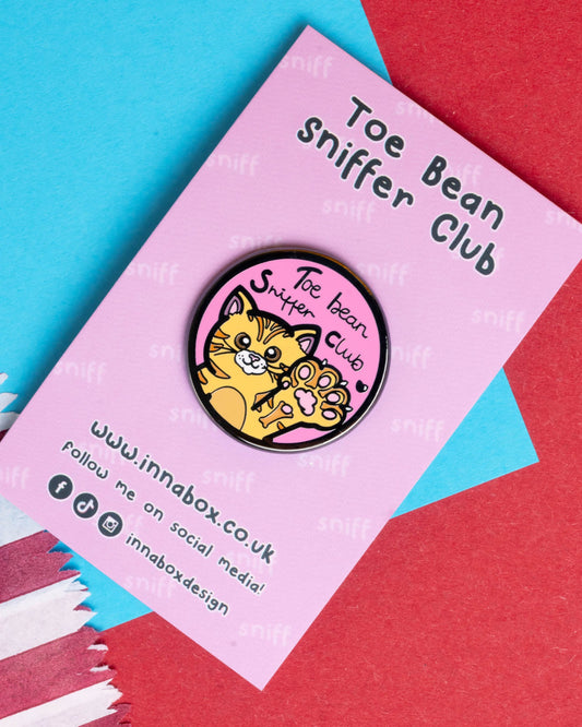 Fun enamel pin badge featuring a cute orange cat holding up a paw, with the phrase "Toe Bean Sniffer Club" written in playful font on a pink circular background. Displayed on branded backing card with pastel pink, blue, and red paper textures. Sold by Rebel Romance, a UK brand celebrating alternative fashion and quirky gifts.