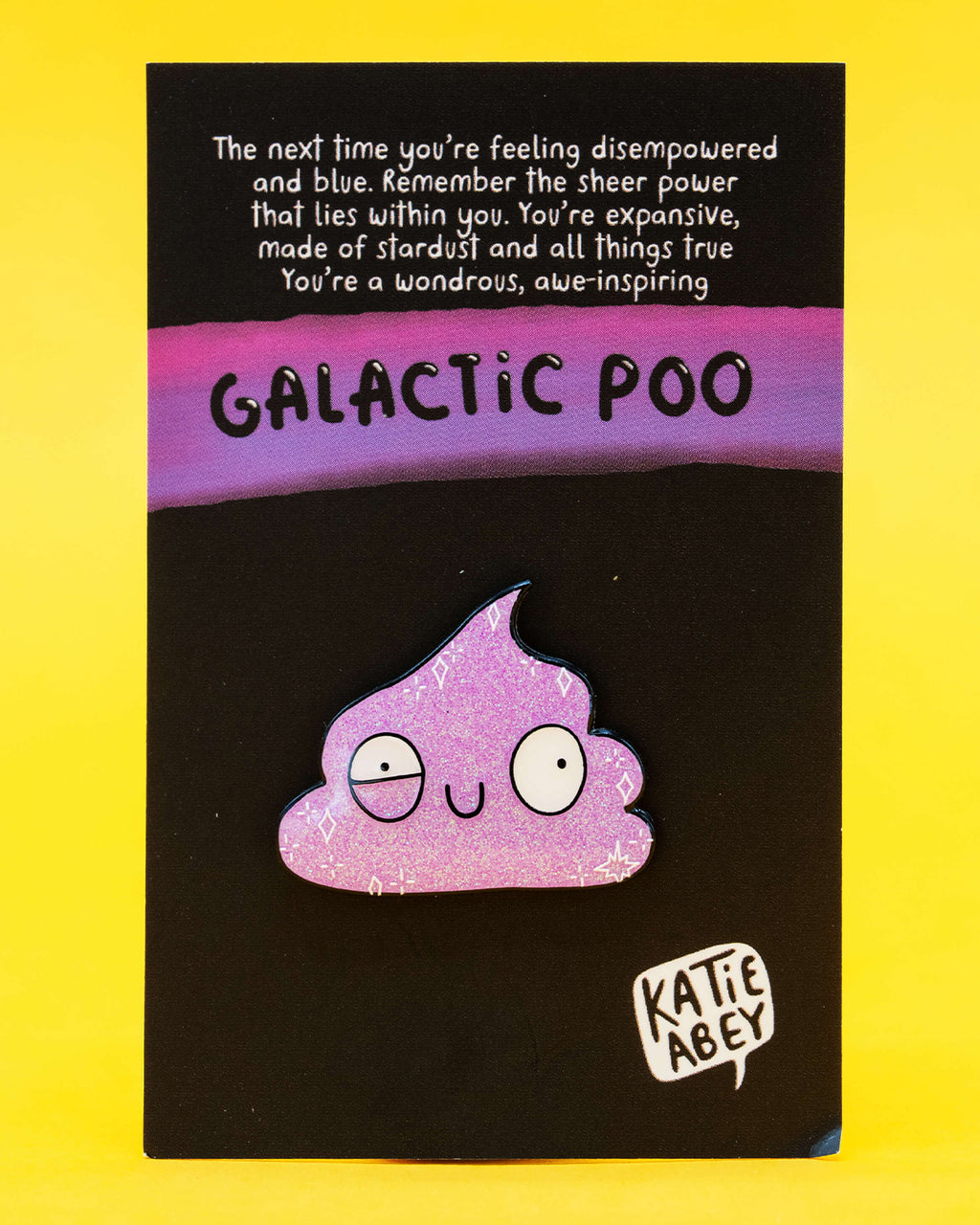 A soft enamel pin displayed on a backing card with a motivational message that reads: “You’re expansive, made of stardust and all things true.” The pin itself is a glittery purple cartoon poo with wide eyes and a silly expression. The card reads “Galactic Poo” and includes the Katie Abey logo. Stocked by Rebel Romance, a UK-based alternative fashion and lifestyle brand.