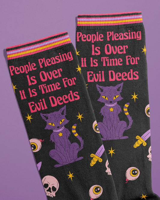 Close-up of the top section of novelty socks featuring the text "People Pleasing Is Over – It Is Time For Evil Deeds" in bold pink above a mystical purple cat. The design includes eyeballs, skulls, knives, and stars on a black background. Designed by FUN CLUB and sold by Rebel Romance, a UK brand embracing extraordinary alternative fashion.