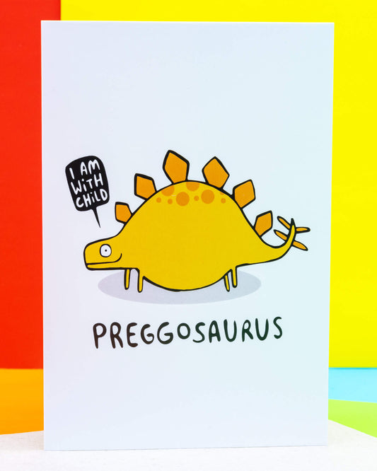A colourful illustrated A6 greeting card from Rebel Romance featuring a pregnant yellow stegosaurus with orange plates and spots, saying “I am with child” in a speech bubble. The text reads “Preggosaurus” in playful black lettering. Set against a bright rainbow background. Designed by UK artist Katie Abey. Ideal for celebrating pregnancy with humour and charm.