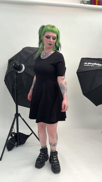 Model posing in Rebel Romance Black Fit & Flare Skater Dress against a white studio background. The soft stretch cotton dress has a flattering fitted top and flared skirt. Styled with chunky platform trainers, highlighting an alternative minimalist gothic fashion look from a UK brand.
