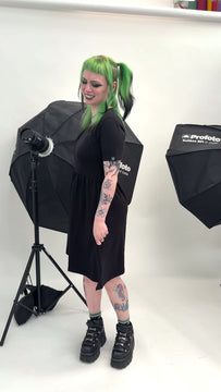 Alternative fashion model with green and black hair wearing the Rebel Romance Black T-Shirt Smock Stretch Dress with Pockets in a white photography studio. Black short sleeve smock dress with a relaxed fit, soft stretch fabric and hidden side pockets, styled with chunky platform boots and tattoos for a gothic casual look.