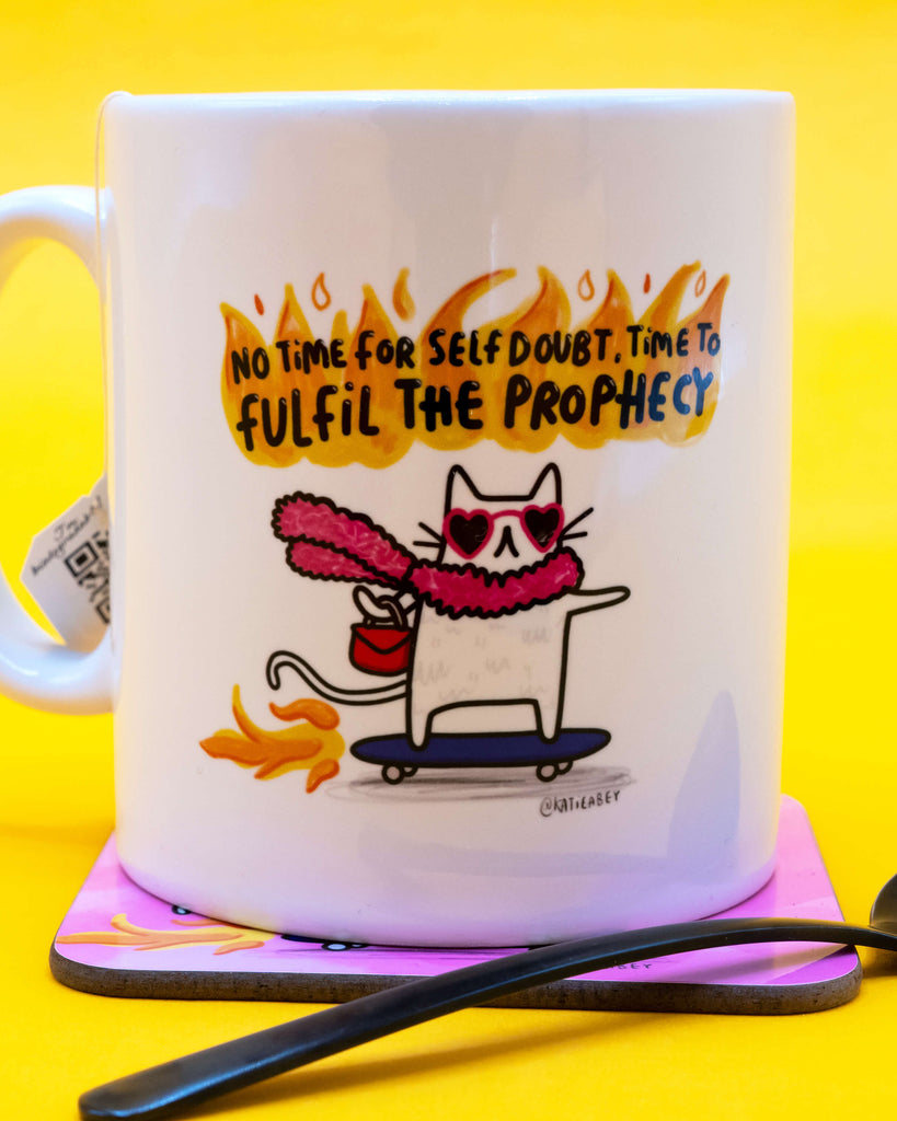Close-up side view of the Time To Fulfil The Prophecy Cat Mug by Katie Abey on a coaster with matching print. Fun and empowering homeware item from Rebel Romance, a UK small business known for celebrating alternative style and creativity.