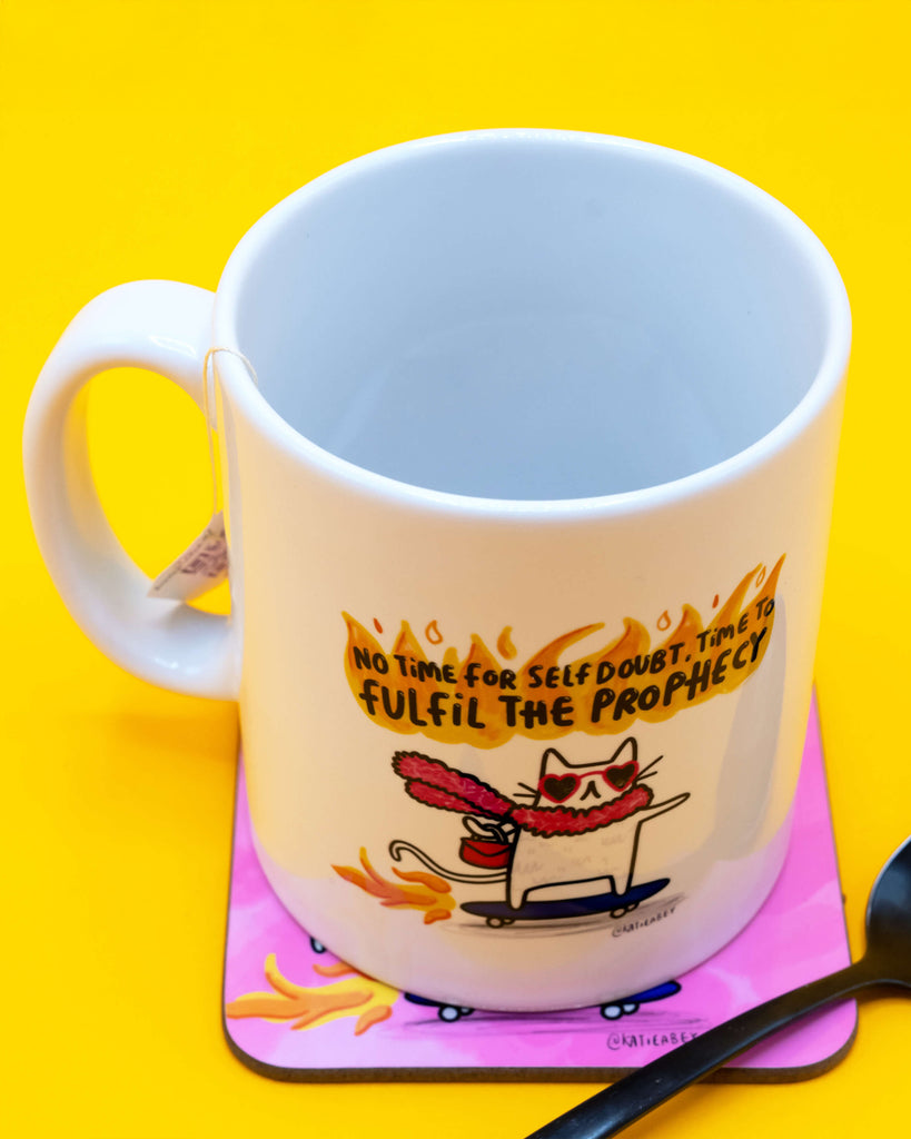 White ceramic mug featuring Katie Abey’s fiery motivational cat design, set against a bold yellow backdrop. The quote reads “No time for self doubt. Time to fulfil the prophecy.” Available at Rebel Romance, a UK indie business selling quirky, uplifting gifts.