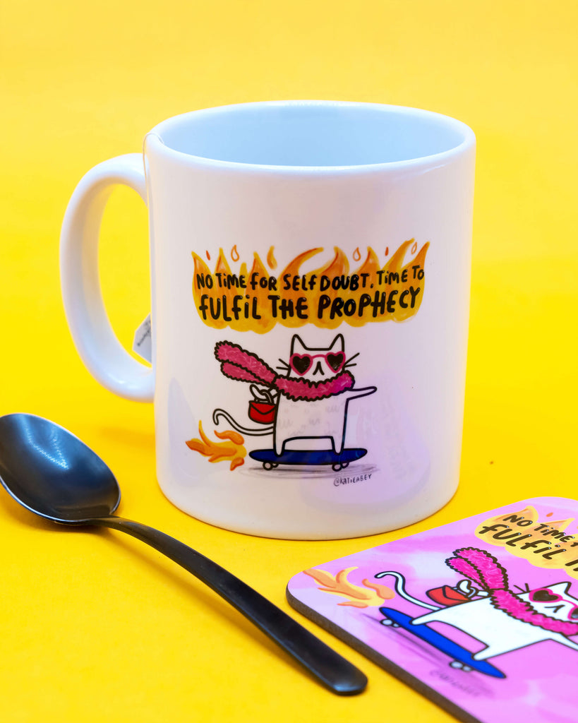 Ceramic mug with a pink coaster and a black spoon, featuring a confident cartoon cat in flames with the quote “No time for self doubt. Time to fulfil the prophecy”. Designed by Katie Abey and sold by Rebel Romance, a UK brand for quirky alternative gifts and homeware.
