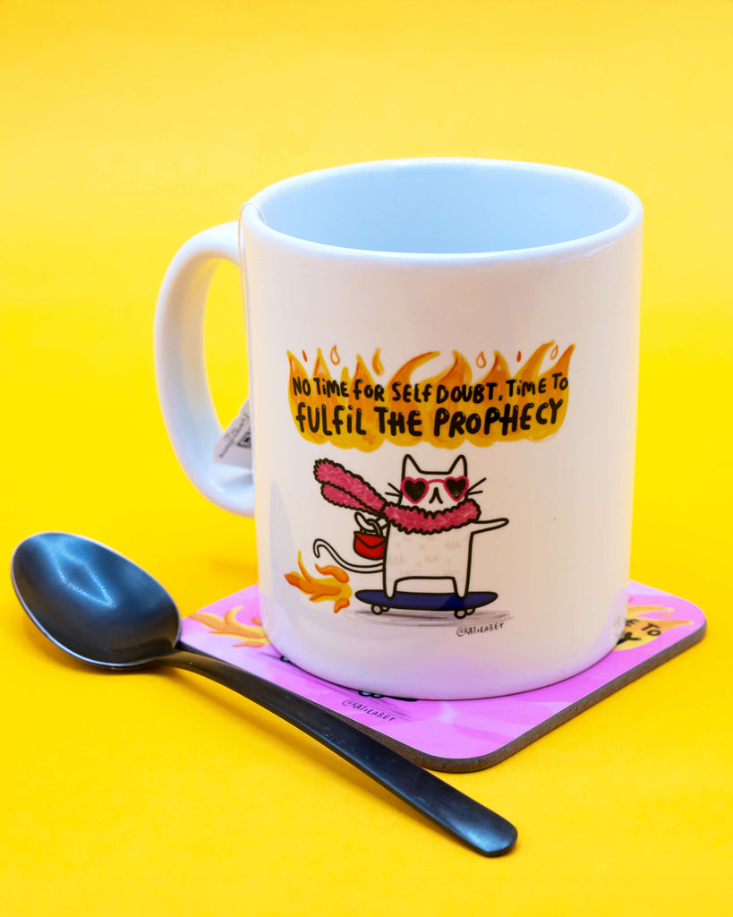 Time To Fulfil The Prophecy Cat Mug by Katie Abey pictured on a pink coaster with matching design, alongside a spoon on a vibrant yellow background. Sold by Rebel Romance, a small UK brand embracing bold, alternative aesthetics and empowering gifts for millennials.