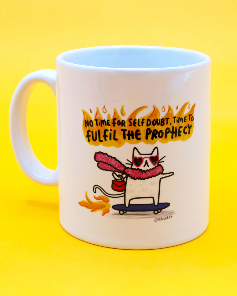 White ceramic mug with colourful illustration of a cat wearing heart sunglasses and a pink fluffy scarf, riding a skateboard through flames with the quote "No time for self doubt. Time to fulfil the prophecy" above in fiery text. Designed by Katie Abey, sold by Rebel Romance, a quirky UK alternative lifestyle brand.
