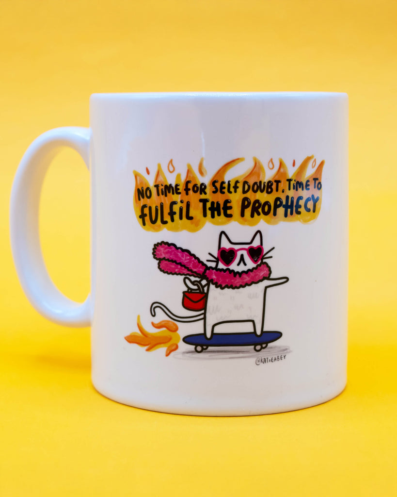 Close-up of a white ceramic mug showing a cat in sunglasses, a pink scarf, and flames, with motivational quote "No time for self doubt. Time to fulfil the prophecy". From Rebel Romance, a UK brand celebrating alternative fashion and empowering homeware with a playful twist.