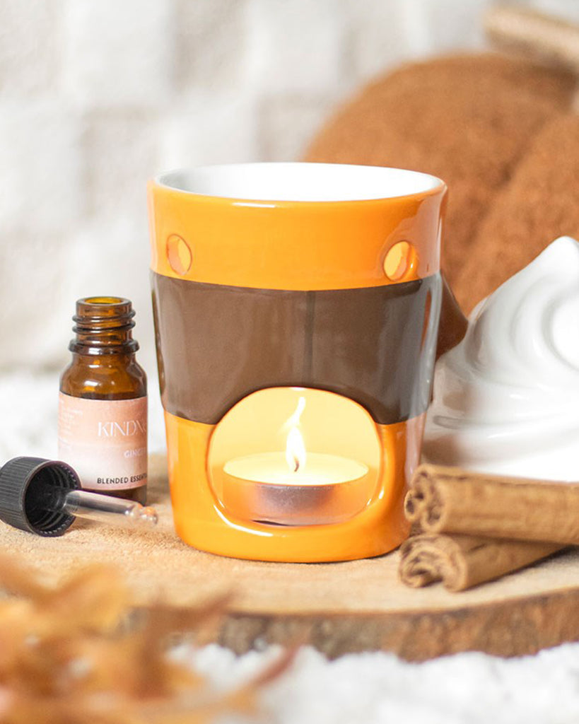 Ceramic pumpkin spice latte oil burner by Rebel Romance in use with a lit tealight inside, lid removed. Autumnal props like cinnamon sticks and essential oil bottles surround it.