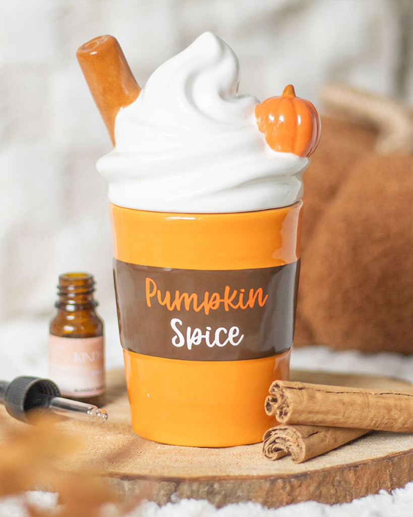 Cosy autumn scene featuring the Rebel Romance ceramic oil burner shaped like a pumpkin spice latte, with whipped cream-style lid, cinnamon stick, and mini pumpkin. "Pumpkin Spice" text is shown on the mug body.