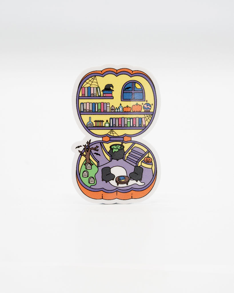 Polly Pocket-inspired die-cut vinyl sticker shaped like an open pumpkin, revealing a spooky miniature library and haunted living room. Features bookshelves, pumpkins, cauldrons, gravestones, and gothic décor in pastel Halloween tones. Designed by Rebel Romance, a small UK brand celebrating alternative fashion, retro nostalgia, and quirky, millennial-friendly spooky gifts.