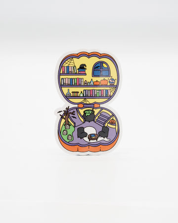 Polly Pocket-inspired die-cut vinyl sticker shaped like an open pumpkin, revealing a spooky miniature library and haunted living room. Features bookshelves, pumpkins, cauldrons, gravestones, and gothic décor in pastel Halloween tones. Designed by Rebel Romance, a small UK brand celebrating alternative fashion, retro nostalgia, and quirky, millennial-friendly spooky gifts.